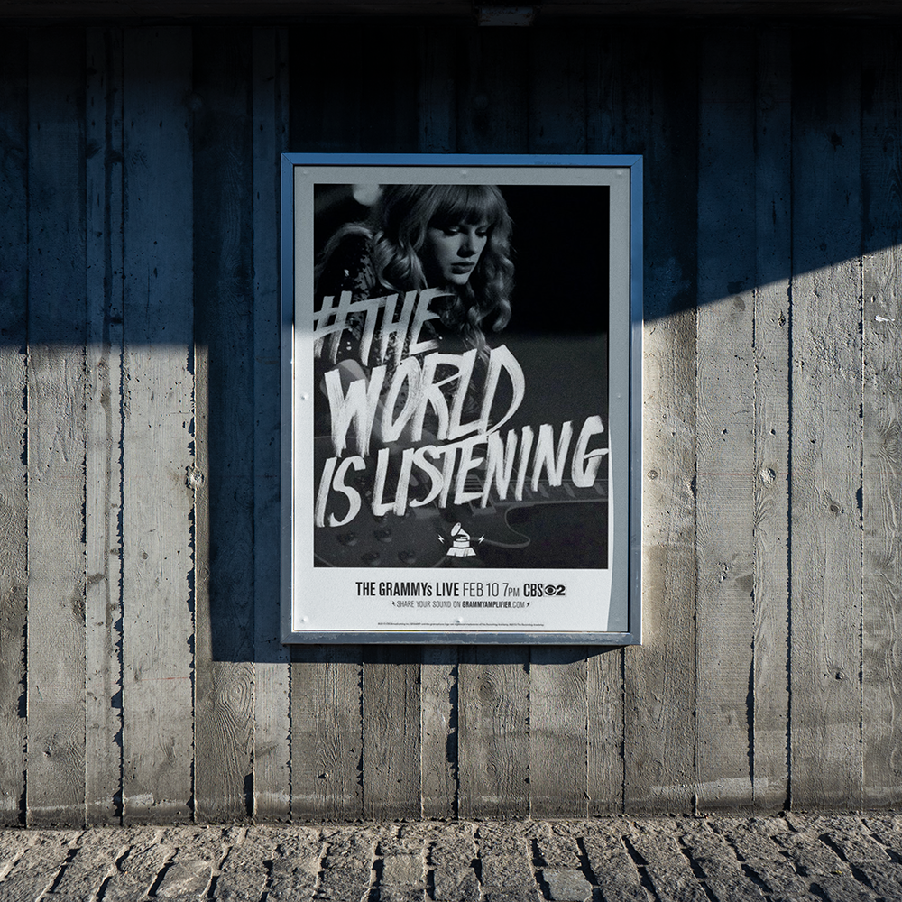 A large poster on a concrete wall features a person with wavy hair holding a microphone. The text on the poster reads, "THE WORLD IS LISTENING" and advertises "The Grammy's Live Feb 10 7PM CBS." The shadow of a railing is cast onto the wall.