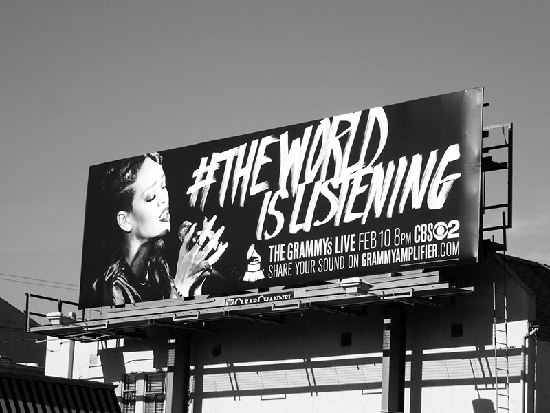 A large billboard displays an image of a musician singing into a microphone with the text "#THEWORLDISLISTENING." Below the main text, additional details include: "THE GRAMMY'S LIVE FEB 10 ON CBS" and the Grammy logo.