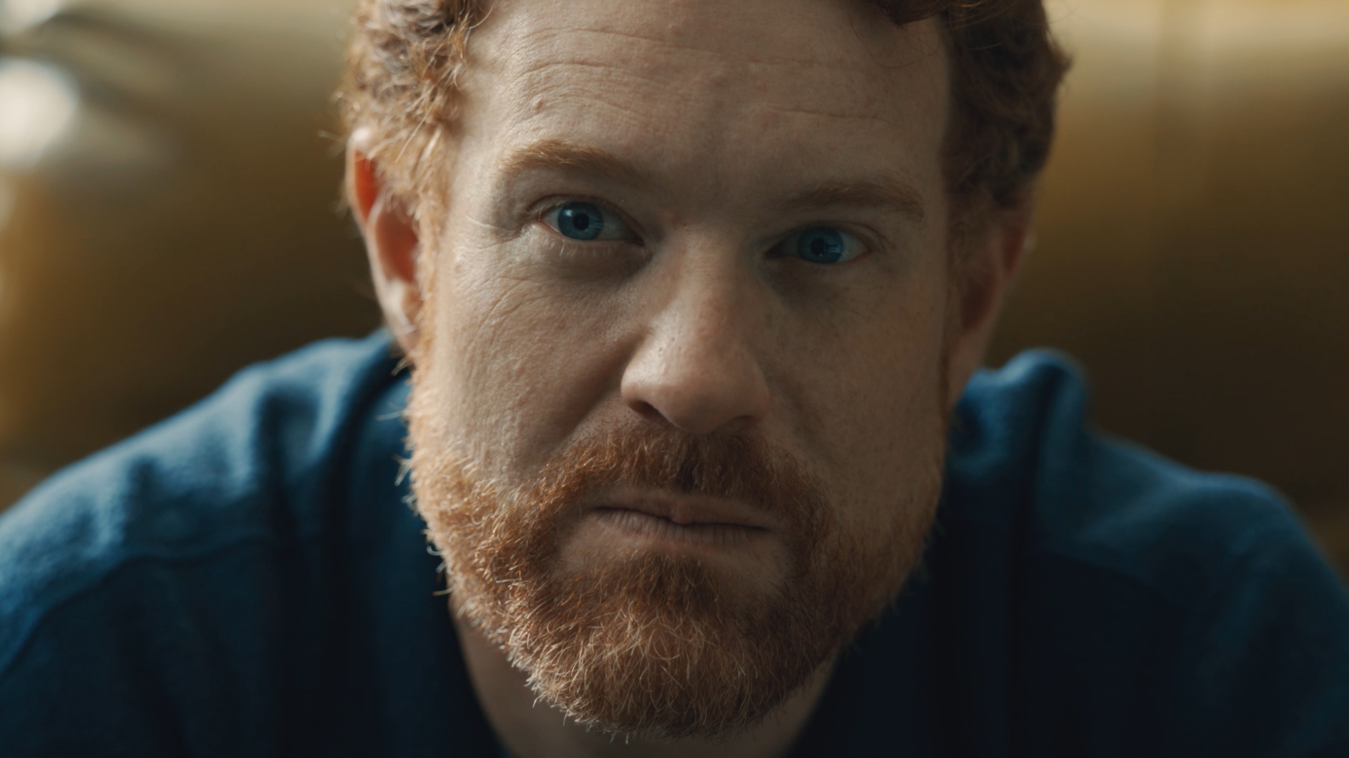 A close-up of a man with short, curly reddish hair and a full beard. He has blue eyes and is wearing a dark shirt. He is looking directly at the camera with a serious expression.