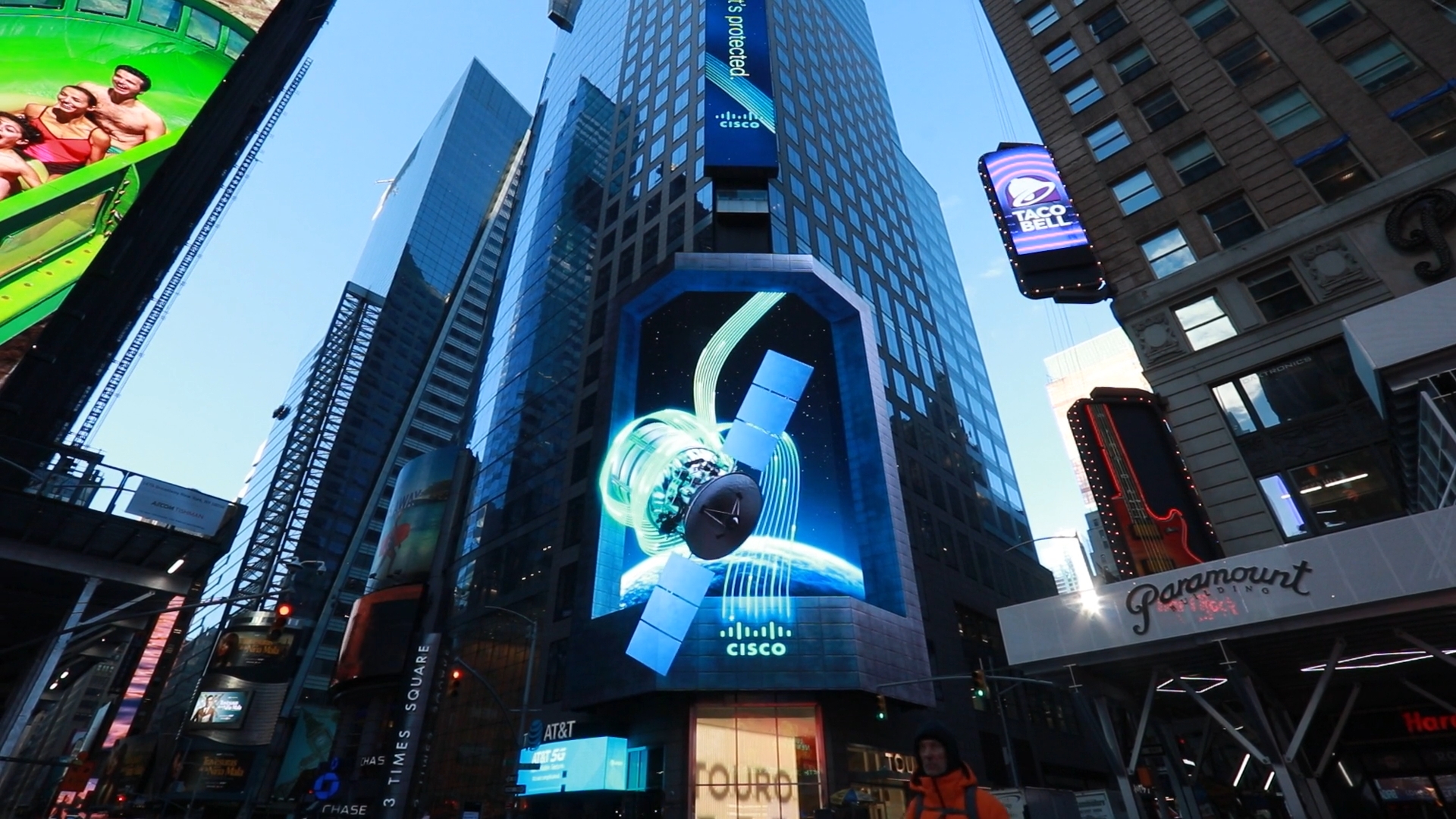 Digital billboards light up Times Square with colorful advertisements on towering skyscrapers. Prominent among them is a large screen showcasing a Cisco ad featuring a satellite in space. Various other ads including Taco Bell and Paramount are also visible.