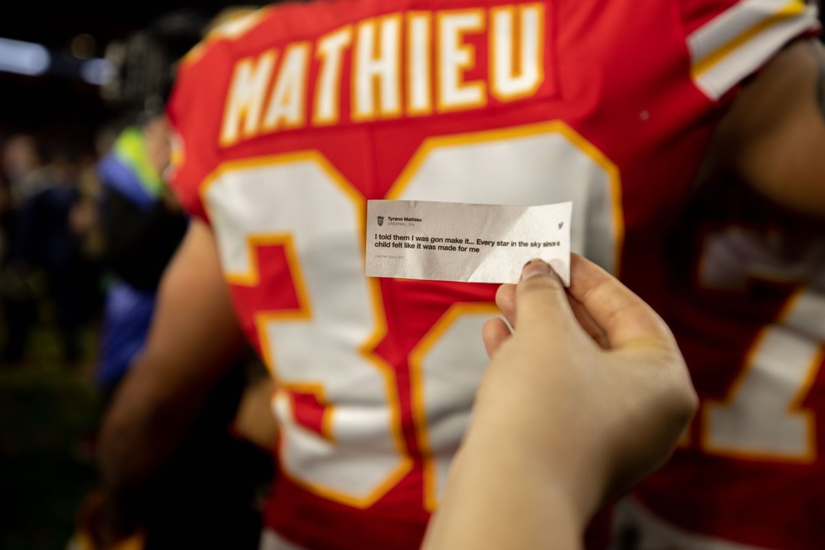 A person's hand holds a piece of paper with a motivational quote, while in the background, a football player wearing a red jersey with yellow accents and the name "Mathieu" on it stands partially visible. The scene appears to be taken on a football field.