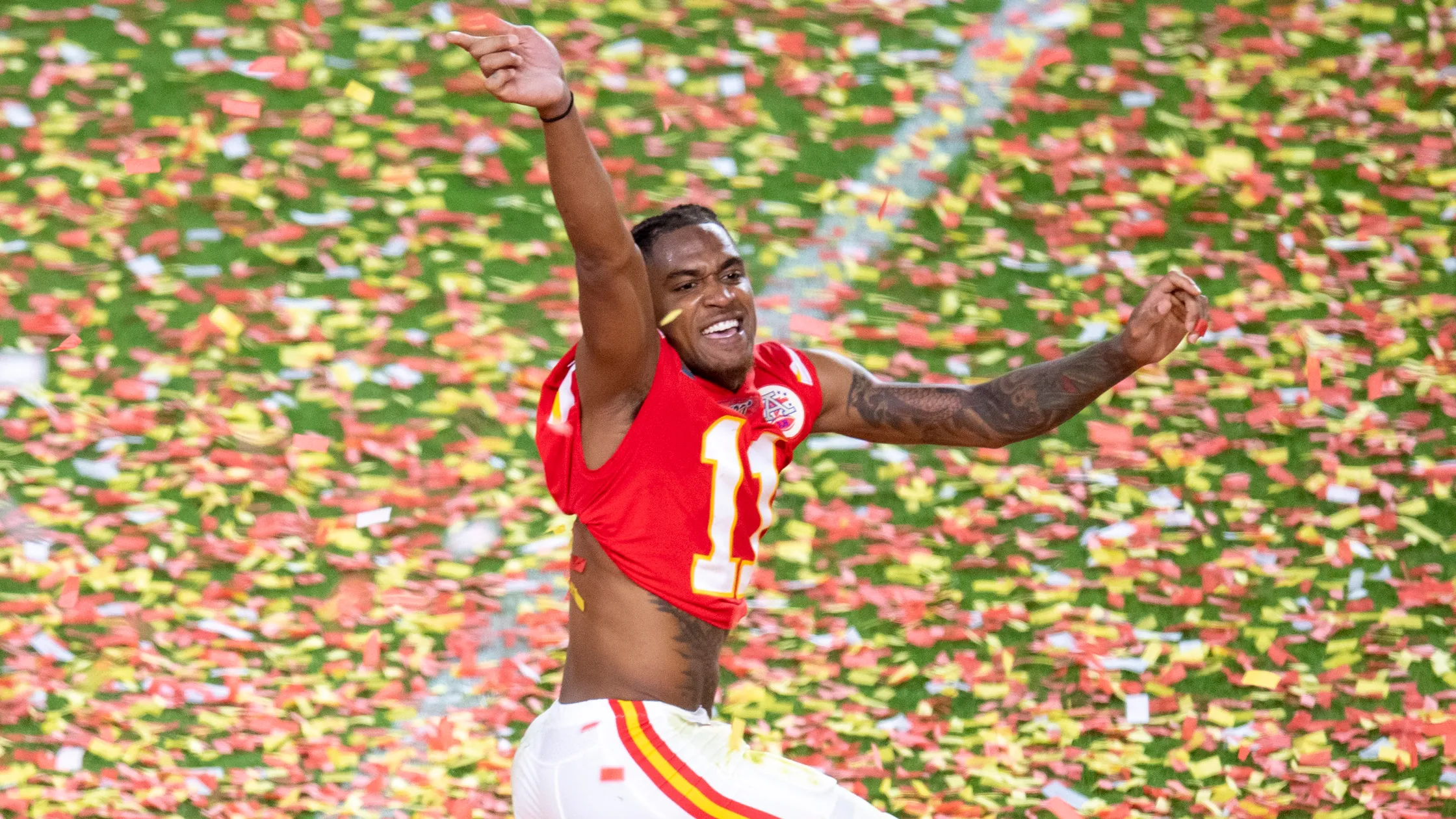 An American football player in a red and white jersey celebrates on a confetti-covered field. He is smiling and has one arm raised while he runs, with colorful confetti falling around him.