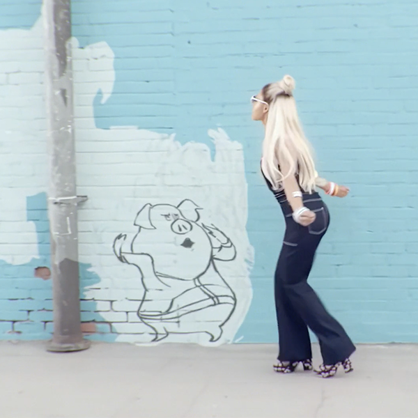 A person with long, blonde hair, wearing sunglasses and flared jeans, poses against a blue brick wall mural. The mural features a dancing pig character. The individual is holding a small round object and appears to be in mid-twirl.