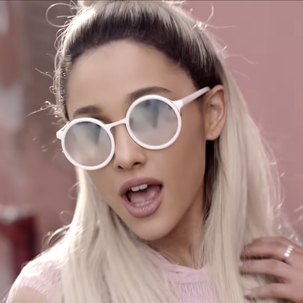 A person with long, blonde hair is shown in close-up wearing large, round, white sunglasses. They are dressed in a light pink top and have their mouth slightly open as if singing, with one hand raised to their shoulder. The background appears to be a blurred outdoor setting.