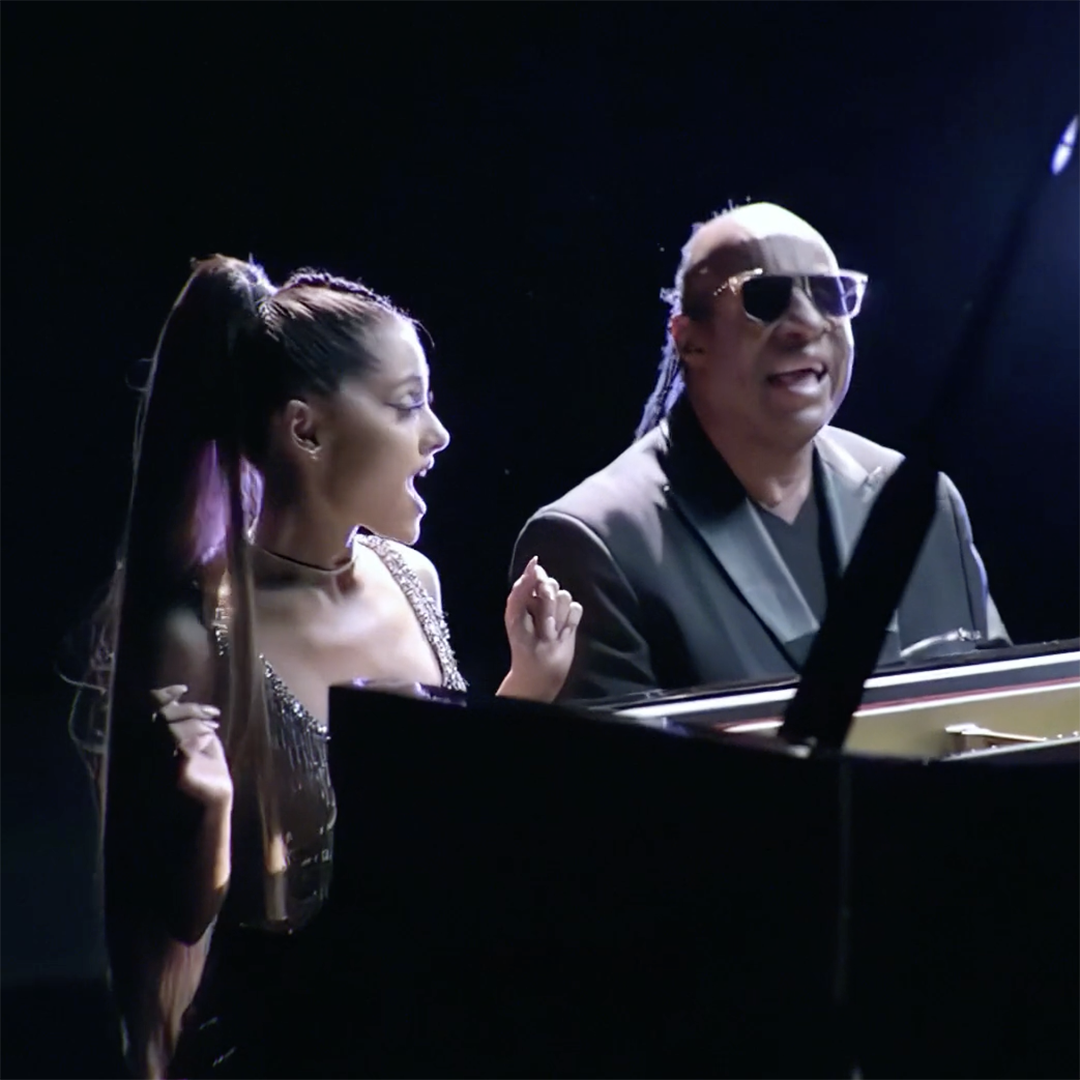 Two individuals are sitting side by side playing a piano. The person on the left has long hair tied in a high ponytail and looks excited. The person on the right is wearing dark glasses and singing. Both appear to be enjoying the music immensely.