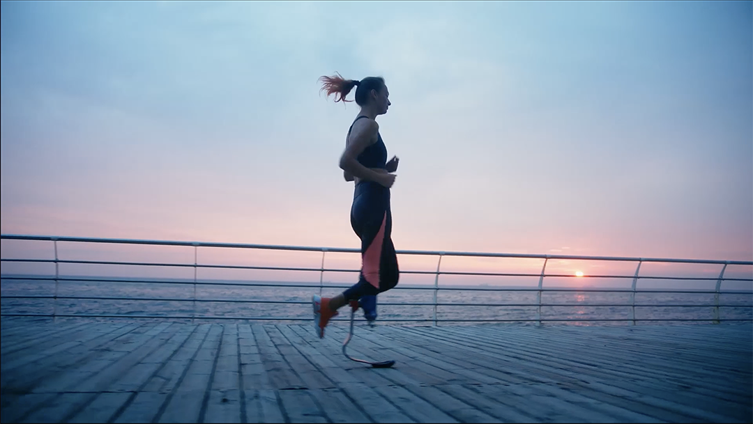 A person with a prosthetic leg runs on a boardwalk by the sea during sunrise or sunset. The sky is filled with a gradient of soft pinks, blues, and purples, creating a serene backdrop. The runner is in motion, showcasing determination and athleticism.