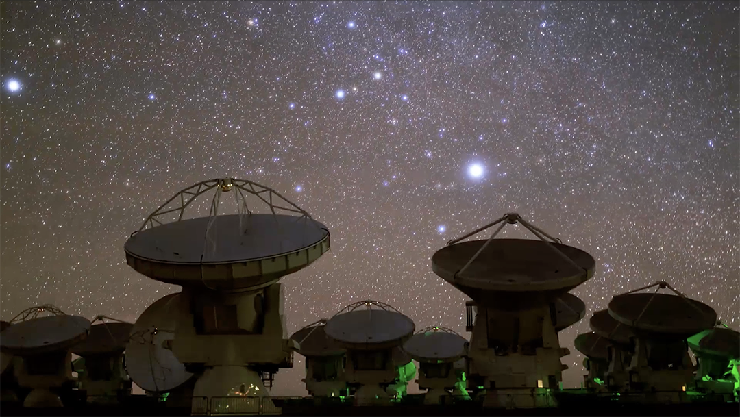 A night view of multiple large radio telescopes with wide dishes, situated under a clear, star-filled sky. The telescopes are illuminated by soft, green lights and a prominent bright star shines in the background.