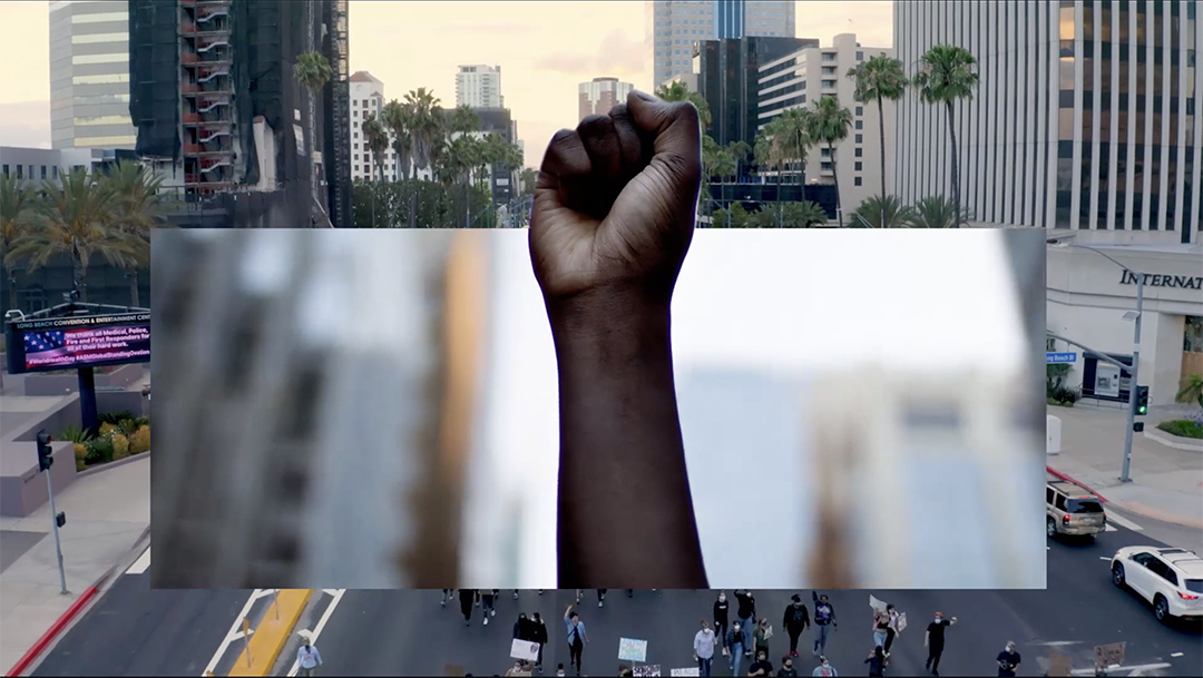 A raised fist is shown in the center of the image, symbolizing solidarity and resistance. The background is a blend of an urban cityscape with tall buildings and a street below with a group of people holding signs, presumably in a protest or march.