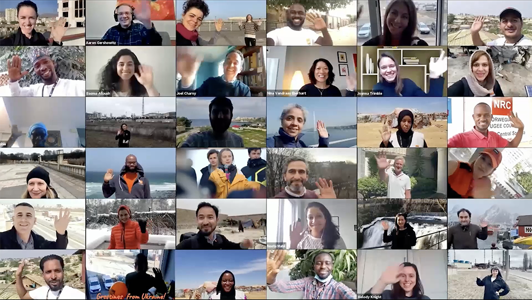 A Zoom screenshot showing a grid of 30 people in various remote locations. Each person is smiling and waving at the camera. The grid features diverse backgrounds, outdoor landscapes, and various personal environments.