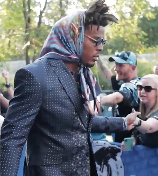A man in a stylish dotted suit and colorful headscarf is seen walking outdoors on a sunny day. Wearing sunglasses, he greets fans reaching out to him. Green trees create a picturesque backdrop.