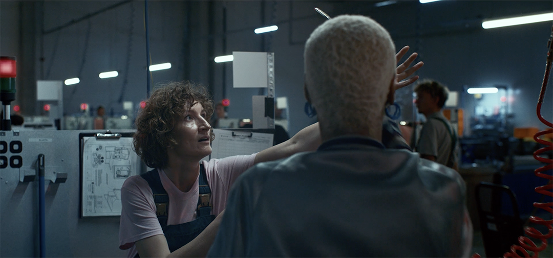 Two people in an industrial setting, engaged in conversation. One has curly hair and is gesturing animatedly. The other, with a short platinum-blonde hairstyle, listens attentively. The background features machinery, workstations, and dim lighting.