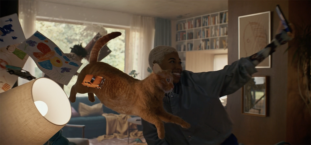 A person wearing a dark shirt is taking a selfie in a cozy living room. Next to them, an orange cat in a brown harness is playfully leaping. The background features a well-lit room with a large window, bookshelves, and children’s drawings.